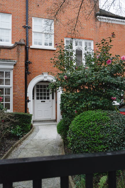 A front garden area in front of a traditional red brick house featuring a white wooden front door with a small window panel, arched door frame, and house number plaque. The garden includes neatly trim